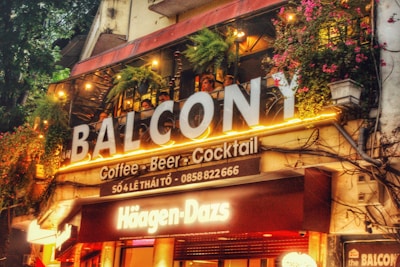 A lively café and bar space with vibrant signs and colorful illumination. The upper balcony is adorned with greenery and hanging lights, creating an inviting atmosphere. The signage prominently features the brands 'Balcôny' and 'Häagen-Dazs'. People are visible enjoying the ambiance on the balcony.