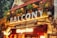 A lively café and bar space with vibrant signs and colorful illumination. The upper balcony is adorned with greenery and hanging lights, creating an inviting atmosphere. The signage prominently features the brands 'Balcôny' and 'Häagen-Dazs'. People are visible enjoying the ambiance on the balcony.