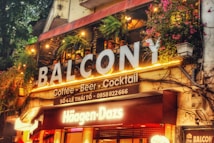A lively caf&eacute; and bar space with vibrant signs and colorful illumination. The upper balcony is adorned with greenery and hanging lights, creating an inviting atmosphere. The signage prominently features the brands 'Balc&ocirc;ny' and 'H&auml;agen-Dazs'. People are visible enjoying the ambiance on the balcony.