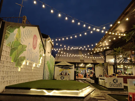 A vibrant outdoor restaurant setting illuminated by strings of lights against a deep blue evening sky. The space features green artificial grass, white walls with plant-themed murals, and a mix of tables and seating areas. Patrons are visible enjoying their meals under large umbrellas.
