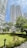 Tall modern residential buildings with curved balconies rise above a well-maintained garden area. Trees and various plants are scattered throughout, providing a lush, green environment. A gazebo sits in the middle of the garden, surrounded by greenery and pathways.