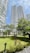 Tall modern residential buildings with curved balconies rise above a well-maintained garden area. Trees and various plants are scattered throughout, providing a lush, green environment. A gazebo sits in the middle of the garden, surrounded by greenery and pathways.