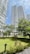 Tall modern residential buildings with curved balconies rise above a well-maintained garden area. Trees and various plants are scattered throughout, providing a lush, green environment. A gazebo sits in the middle of the garden, surrounded by greenery and pathways.