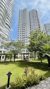 Tall modern residential buildings with curved balconies rise above a well-maintained garden area. Trees and various plants are scattered throughout, providing a lush, green environment. A gazebo sits in the middle of the garden, surrounded by greenery and pathways.