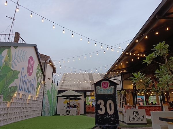 An outdoor dining area featuring string lights hanging above, with vibrant murals on the walls and green artificial grass covering the ground. There are tables with chairs under big umbrellas and some decorative plants. A sign with the number 30 and the logo 'Mie Gacoan' is prominently displayed in the foreground.