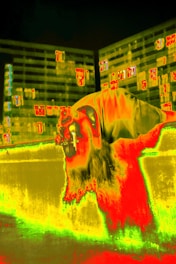 A vibrant thermal image featuring a person leaning over a ledge, possibly using binoculars. The background shows abstract and geometric patterns in bright colors, resembling buildings with windows.
