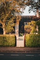 A quaint, charming cottage with a dark gabled roof and white picket gate. Lush green hedges and tall trees frame the entrance. Warm sunlight casts shadows, giving the scene a serene, peaceful atmosphere.