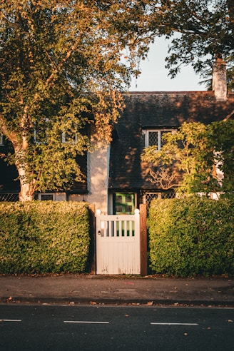 A quaint, charming cottage with a dark gabled roof and white picket gate. Lush green hedges and tall trees frame the entrance. Warm sunlight casts shadows, giving the scene a serene, peaceful atmosphere.