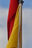 Red and yellow flags attached to a wooden pole, with a background of a cloudy blue sky.
