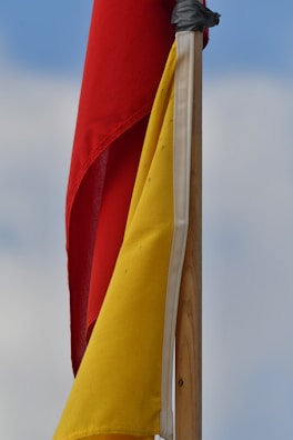 Red and yellow flags attached to a wooden pole, with a background of a cloudy blue sky.