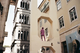 An ornate and intricately designed elevator structure appears alongside a beige building with multiple windows. A striking pink statue of a person in a suit stands prominently on an architectural feature resembling a bridge. Surrounding buildings exhibit a historic and artistic style, and a piece of street art is partially visible on the right.