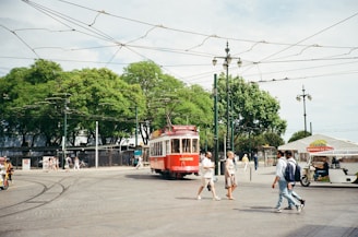 A red tram is moving along tracks in a spacious, open area with large, leafy green trees in the background. Several people are walking nearby, including a couple crossing the street. Overhead, there are multiple wires and streetlights. In the background, there is a vendor stall with the sign 'mbooks MERCADO DO LIVRO'. The sky is clear, suggesting a bright day.