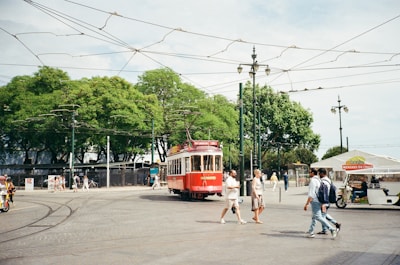 A red tram is moving along tracks in a spacious, open area with large, leafy green trees in the background. Several people are walking nearby, including a couple crossing the street. Overhead, there are multiple wires and streetlights. In the background, there is a vendor stall with the sign 'mbooks MERCADO DO LIVRO'. The sky is clear, suggesting a bright day.