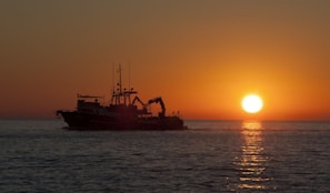A scenic coastal sunset with calm waves and a traditional fishing boat silhouetted against the orange sky.