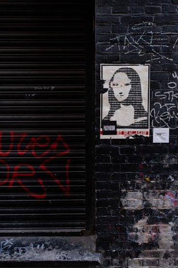 A dark brick wall featuring a poster with a pixelated portrait composed of binary numbers, resembling a famous painting. Red graffiti is spray-painted on the left side of the wall, while white chalk-like scribbles are present around the poster. The wall has a worn, urban look.