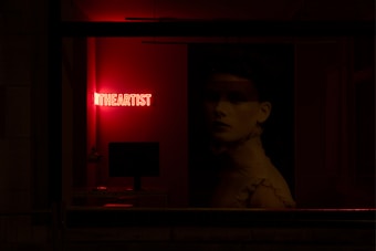 A dimly lit room features a bright red neon sign that reads 'THE ARTIST.' A computer monitor sits on a desk, and there is a large portrait of a woman with a somber expression. The room has an overall dark ambiance with red lighting accents.