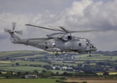 A military helicopter with the Royal Navy insignia is flying over a scenic, rural landscape. The fields below are divided into patches of various shades of green and brown, possibly indicating farmland. The helicopter is grey in color, with a visible rotor in motion.
