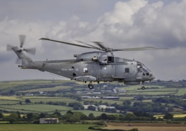 A military helicopter with the Royal Navy insignia is flying over a scenic, rural landscape. The fields below are divided into patches of various shades of green and brown, possibly indicating farmland. The helicopter is grey in color, with a visible rotor in motion.