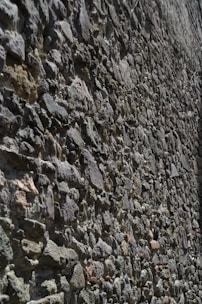 A textured stone wall composed of unevenly arranged rocks of various shapes and sizes. The stones are in earthy tones and are tightly packed together, creating a rough surface with visible grooves and shadows.