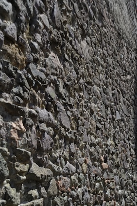 A textured stone wall composed of unevenly arranged rocks of various shapes and sizes. The stones are in earthy tones and are tightly packed together, creating a rough surface with visible grooves and shadows.