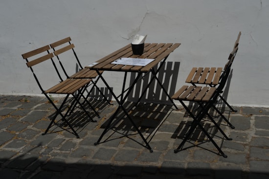 A minimalist outdoor setup with a wooden table and three matching folding chairs placed on a cobblestone pavement. A menu or a piece of paper and a small container, possibly for napkins, rest on the table. The area is brightly lit by sunlight, creating distinct shadows on the ground and against the plain white wall backdrop.