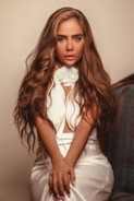 A person with long, wavy brown hair is seated, wearing a white outfit with a floral detail around the neck and a pearl necklace. The background is softly blurred, focusing attention on the person, who exudes confidence and glamour.
