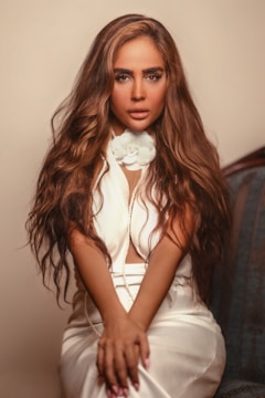 A person with long, wavy brown hair is seated, wearing a white outfit with a floral detail around the neck and a pearl necklace. The background is softly blurred, focusing attention on the person, who exudes confidence and glamour.
