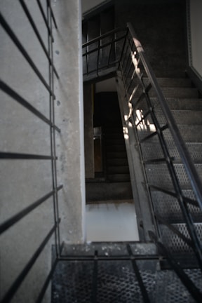 A staircase with metal railings and grated steps is viewed from an angle, creating a geometric play of lines and shadows. The stairs ascend and descend, leading to different levels. The surrounding walls are concrete, with a textured and slightly worn appearance, adding to the industrial aesthetic.