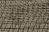 A grid of steel reinforcing bars casts intricate shadows on a rough concrete surface, creating a pattern of intersecting lines. The bars have a rusty appearance, contributing to the industrial aesthetic.