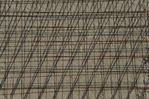A grid of steel reinforcing bars casts intricate shadows on a rough concrete surface, creating a pattern of intersecting lines. The bars have a rusty appearance, contributing to the industrial aesthetic.