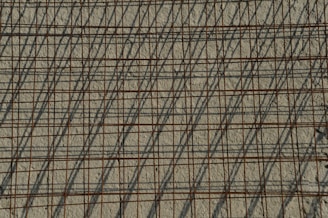A grid of steel reinforcing bars casts intricate shadows on a rough concrete surface, creating a pattern of intersecting lines. The bars have a rusty appearance, contributing to the industrial aesthetic.