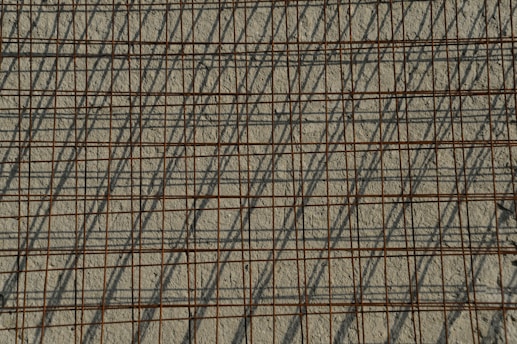 A grid of steel reinforcing bars casts intricate shadows on a rough concrete surface, creating a pattern of intersecting lines. The bars have a rusty appearance, contributing to the industrial aesthetic.