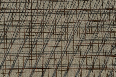 A grid of steel reinforcing bars casts intricate shadows on a rough concrete surface, creating a pattern of intersecting lines. The bars have a rusty appearance, contributing to the industrial aesthetic.
