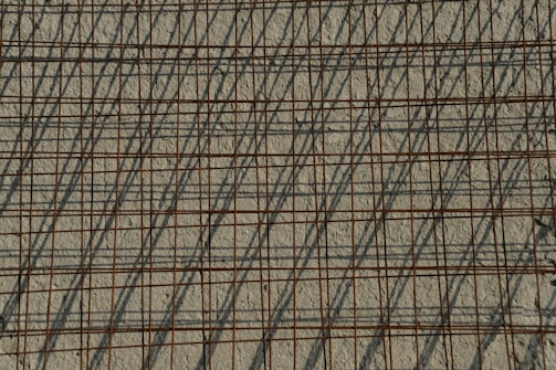 A grid of steel reinforcing bars casts intricate shadows on a rough concrete surface, creating a pattern of intersecting lines. The bars have a rusty appearance, contributing to the industrial aesthetic.