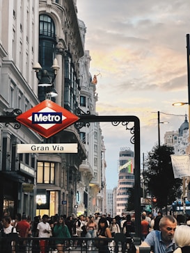 A bustling city street scene featuring a metro sign for Gran V&iacute;a, surrounded by historic buildings with detailed architecture. The sky above is a mix of clouds with hints of sunset colors, creating a dramatic backdrop. A large crowd of people can be seen walking along the street, adding to the lively atmosphere of the area.
