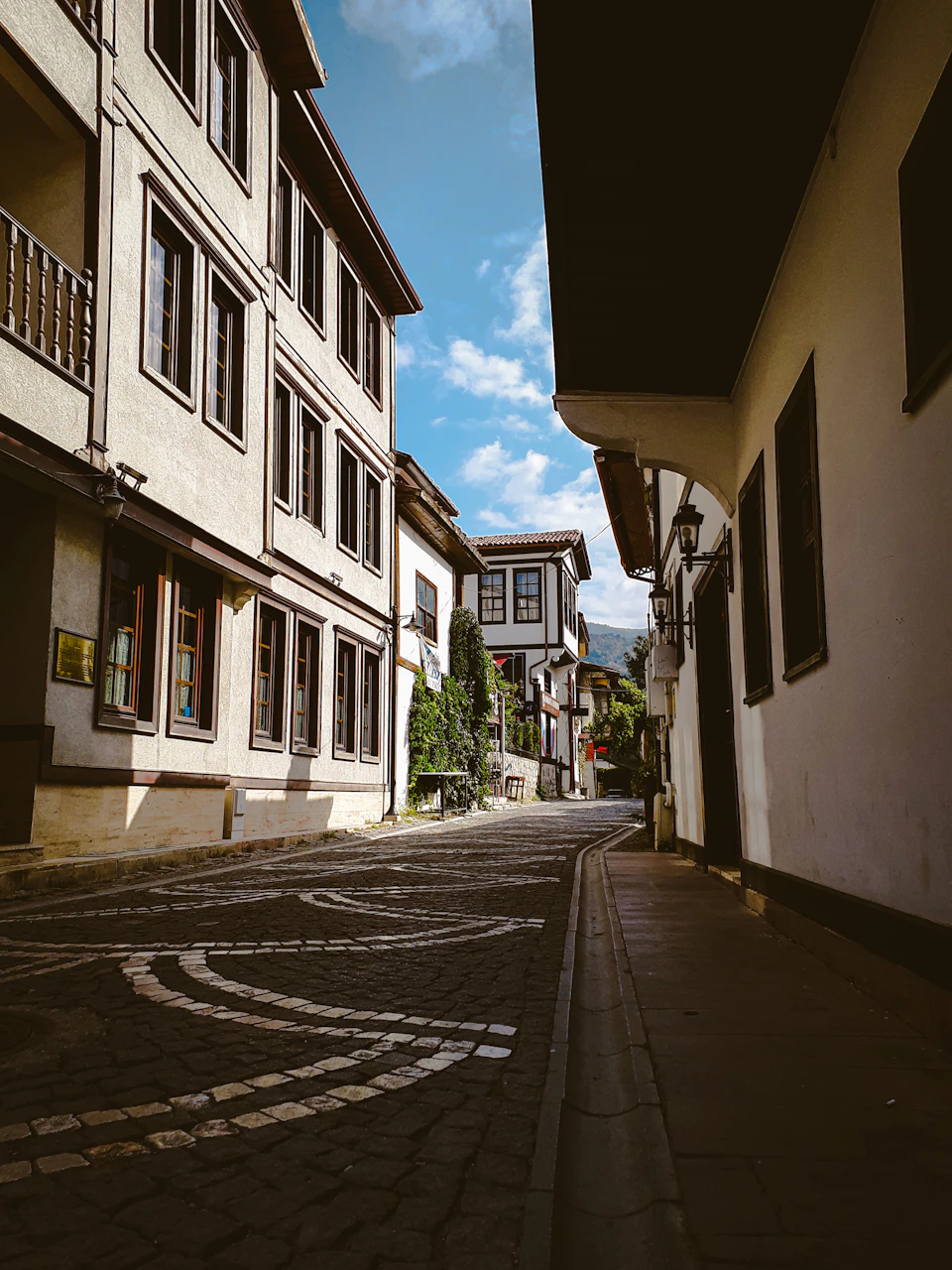 The image depicts a narrow cobblestone street flanked by old, white buildings with balconies on both sides. The buildings have dark wooden shutters and are painted white, creating a contrast against the clear blue sky above. The perspective is from ground level, looking down the street towards the horizon line, giving a sense of depth to the scene.