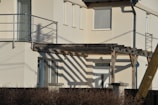 A modern building facade featuring light beige walls with large glass windows. There is a wooden pergola casting distinct shadows, accompanied by a metal railing balcony above.