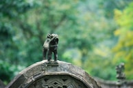 A small, intricate statue of a lion stands on top of an ornate stone structure, surrounded by a lush, green forest. The lion appears to be roaring or expressing a powerful stance.