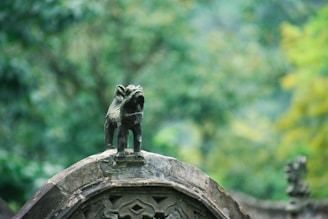 A small, intricate statue of a lion stands on top of an ornate stone structure, surrounded by a lush, green forest. The lion appears to be roaring or expressing a powerful stance.