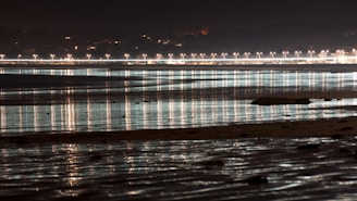A nighttime coastal scene featuring a series of bright streetlights lining a distant shoreline. The lights reflect off the calm water, creating long streaks of illumination. The sky is dark, contrasting with the warm glow of the streetlights.