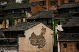An aged building facade features a map of China created using varied shades of bricks set against a worn-out, plastered wall. The surrounding structures exhibit traditional Chinese architecture with dark, tiled roofs and weathered bricks, partially covered by climbing green plants.