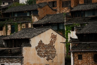 An aged building facade features a map of China created using varied shades of bricks set against a worn-out, plastered wall. The surrounding structures exhibit traditional Chinese architecture with dark, tiled roofs and weathered bricks, partially covered by climbing green plants.