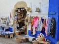 A small storefront displaying various items for sale, including bags, aprons, and colorful textiles. Baskets filled with hats and other small items are placed on the ground. The exterior wall is painted blue and white, adding a vibrant backdrop to the display.