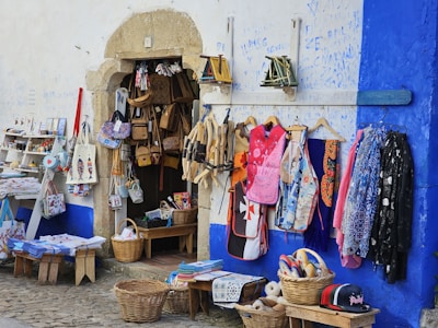 A small storefront displaying various items for sale, including bags, aprons, and colorful textiles. Baskets filled with hats and other small items are placed on the ground. The exterior wall is painted blue and white, adding a vibrant backdrop to the display.
