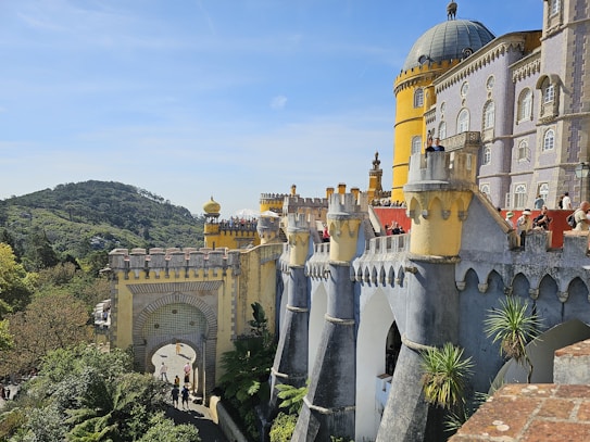 A colorful palace with intricate architecture featuring towers and arched structures. The facade displays a mix of yellow and purple colors, with decorative details. The backdrop includes lush greenery and a clear blue sky. Several people are present, admiring the view.