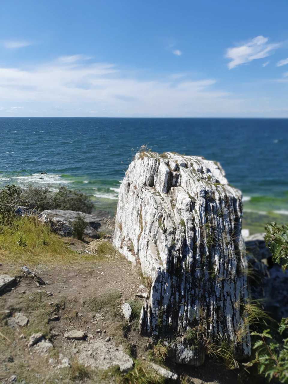 The image depicts a rocky cliff overlooking the ocean, with a large white rock formation on the right side and a smaller one on the left. The cliff is covered in green grass and shrubs, adding a touch of nature to the scene. In the background, the vast expanse of the ocean stretches out towards the horizon under a clear blue sky dotted with fluffy white clouds.