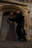 Two people stand confidently in front of a large, ornate wooden door framed by detailed stonework. One person is dressed in a long, fitted brown dress with hands on the hips, while the other wears all black with oversized sunglasses and dramatic platform shoes. The scene conveys a sense of boldness and style.