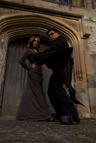 Two people stand confidently in front of a large, ornate wooden door framed by detailed stonework. One person is dressed in a long, fitted brown dress with hands on the hips, while the other wears all black with oversized sunglasses and dramatic platform shoes. The scene conveys a sense of boldness and style.