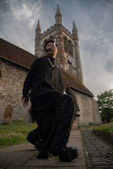 A person dressed in dark clothing with unique and futuristic fashion elements poses in front of an old stone church with tall spires and a clock on its facade. The scene looks dramatic with cloudy skies in the background, creating a strong contrast between the modern attire and the historic architecture.