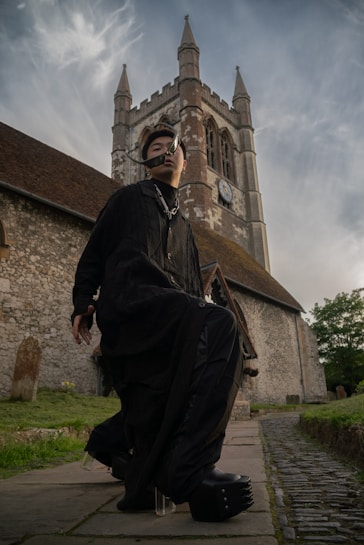 A person dressed in dark clothing with unique and futuristic fashion elements poses in front of an old stone church with tall spires and a clock on its facade. The scene looks dramatic with cloudy skies in the background, creating a strong contrast between the modern attire and the historic architecture.
